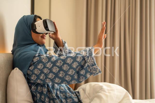 التطور التقني والتكنولوجي, المتعة و التسلية بالمشاهدة عبر نظارة الواقع الافتراضي،  إمرأة عربية خليجية سعودية محجبة ترتدي العباءة تجلس على السرير في المنزل وتنظر من خلال نظارات ثلاثية الأبعاد، الاندماج ومحاكاة العالم الخيالي ، ايماءات السعادة والسرور