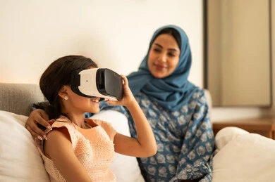 Fun and entertainment watching through virtual reality glasses, a Saudi Arabian Gulf girl wearing a dress sitting on the bed at home with her mother and looking through 3D glasses, merging and simulating the imaginary world, gestures of happiness and pleasure, technical and technological development