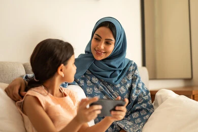 A Saudi Arabian Gulf girl wearing a dress holding a mobile phone, using a modern and advanced technical device, a mother wearing an abaya sitting with her daughter on the bed, the concept of e-learning, following lessons remotely via the Internet, educational scientific curricula