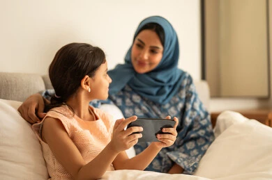   فتاة عربية خليجية سعودية ترتدي الفستان تحمل بيدها الهاتف المحمول، استخدام جهاز تقني حديث ومتطور، ام ترتدي العباءة تجلس مع ابتنها على السرير، مفهوم التعليم الالكتروني، متابعة الدروس عن بعد عن طريق الانترنت، المناهج العلمية التعليمية