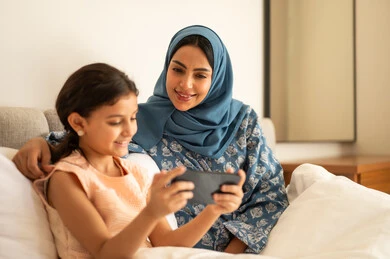   فتاة عربية خليجية سعودية ترتدي الفستان تحمل بيدها الهاتف المحمول، استخدام جهاز تقني حديث ومتطور، ام ترتدي العباءة تجلس مع ابتنها على السرير، مفهوم التعليم الالكتروني، متابعة الدروس عن بعد عن طريق الانترنت، المناهج العلمية التعليمية