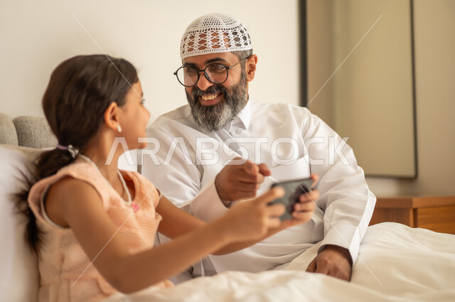 اب عربي خليجي سعودي يرتدي الثوب التقليدي والقبعة البيضاء يجلس على السرير مع ابنته،  فتاة عربية خليجية سعودية تحمل بيدها الهاتف المحمول، استخدام جهاز تقني حديث ومتطور، مفهوم التعليم الالكتروني، متابعة الدروس عن بعد عن طريق الانترنت، المناهج العلمية التعليمية