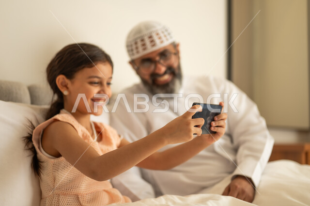 اب عربي خليجي سعودي يرتدي الثوب التقليدي والقبعة البيضاء يجلس على السرير مع ابنته،  فتاة عربية خليجية سعودية تحمل بيدها الهاتف المحمول، استخدام جهاز تقني حديث ومتطور، مفهوم التعليم الالكتروني، متابعة الدروس عن بعد عن طريق الانترنت، المناهج العلمية التعليمية