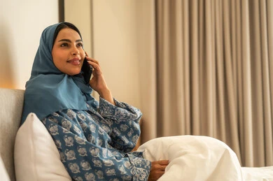 Chatting with family and friends, browsing social media apps and programs, working remotely, a smiling veiled Saudi Arabian Gulf woman wearing an abaya sitting on the bed holding a mobile phone in her hand, using modern and advanced  devices
