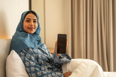 عرض شاشة سوداء فارغة عبر الجوال، الدردشه مع الأهل والأصدقاء، تصفح تطبيقات وبرامج التواصل الاجتماعي، العمل عن بعد، سيدة عربية خليجية سعودية محجبة مبتسمة ترتدي عباءة تجلس على السرير تمسك بيدها الهاتف المحمول، استخدام الاجهزة التقنية الحديثة والمتطورة