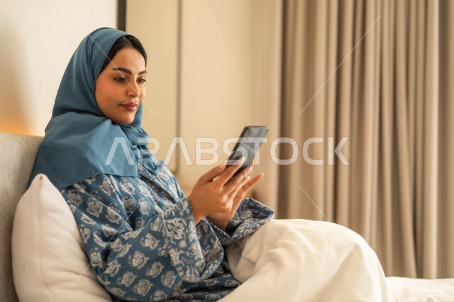 Chatting with family and friends, browsing social media apps and programs, working remotely, a smiling veiled Saudi Arabian Gulf woman wearing an abaya sitting on the bed holding a mobile phone in her hand, using modern and advanced technical devices