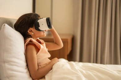Technical and technological development, pleasure and entertainment by watching via virtual reality glasses, Arab Gulf girl, wearing the dress sitting on the bed at home and looking through three -dimensional glasses, merger and simulation of the imaginary world, gestures of happiness and pleasure