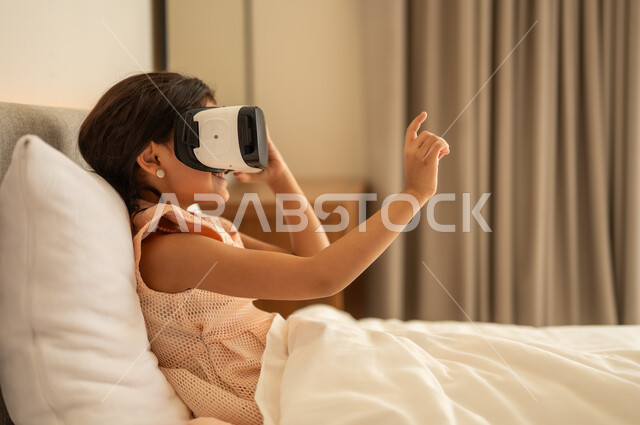 التطور التقني والتكنولوجي, المتعة و التسلية بالمشاهدة عبر نظارة الواقع الافتراضي،  فتاة عربية خليجية سعودية ترتدي الفستان تجلس على السرير في المنزل وتنظر من خلال نظارات ثلاثية الأبعاد، الاندماج ومحاكاة العالم الخيالي ، ايماءات السعادة والسرور