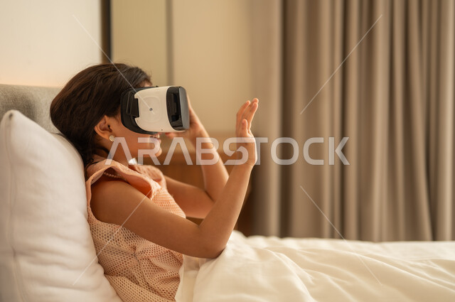 Technical and technological development, pleasure and entertainment by watching via virtual reality glasses, Arab Gulf girl, wearing the dress sitting on the bed at home and looking through three -dimensional glasses, merger and simulation of the imaginary world, gestures of happiness and pleasure