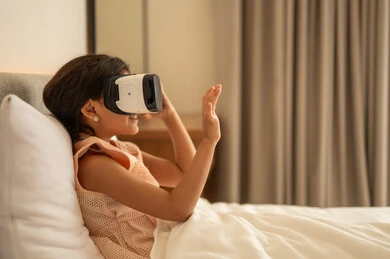 Technical and technological development, pleasure and entertainment by watching via virtual reality glasses, Arab Gulf girl, wearing the dress sitting on the bed at home and looking through three -dimensional glasses, merger and simulation of the imaginary world, gestures of happiness and pleasure