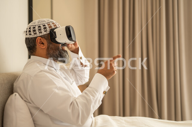 التفاعل والاندماج ومحاكاة العالم الخيالي، رجل عربي خليجي سعودي مبتسم يرتدي الثوب التقليدي والقبعة البيضاء بجلس على السرير يضع نظارة الواقع الافتراضي، المتعة والتسلية بالمشاهدة عبر نظارات ثلاثية الابعاد
