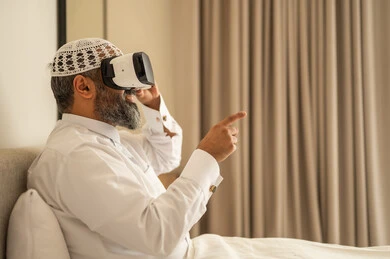 Interaction, integration and simulation of the imaginary world, a smiling Saudi Arabian Gulf man wearing traditional clothes and a white hat sitting on the bed wearing virtual reality glasses, fun and entertainment watching through 3D glasses
