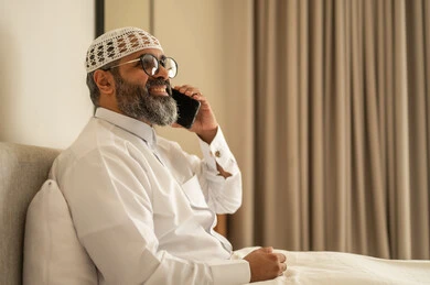 تصفح مواقع التواصل الاجتماعي، رجل عربي خليجي سعودي يرتدي الثوب التقليدي والقبعة البيضاء يجلس على السرير يمسك بيده الهاتف المحمول، الدردشة والتواصل مع الأهل و الأصدقاء، استخدام جهاز تقني حديث ومتطور، العمل عن بعد وانجاز المهام المطلوبة