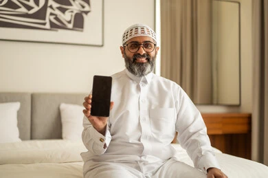 عرض شاشة سوداء فارغة، تصفح مواقع التواصل الاجتماعي، رجل عربي خليجي سعودي يرتدي الثوب التقليدي والقبعة البيضاء يجلس على السرير يمسك بيده الهاتف المحمول، الدردشة والتواصل مع الأهل و الأصدقاء، استخدام جهاز تقني حديث ومتطور، العمل عن بعد وانجاز المهام المطلوبة