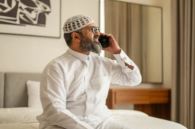 Browsing social media, a Saudi Arabian Gulf man wearing traditional thobe and white hat sitting on the bed holding a mobile phone, chatting and communicating with family and friends, using a modern and advanced technical device, working remotely and coleting the required tasks