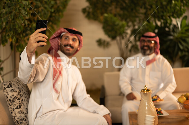 Taking a selfie with your mobile phone, chatting, a happy family gathering, expressions of affection and love, enjoying time with your children, an elderly Saudi Arabian Gulf man wearing a traditional thobe and shemagh sitting on the sofa next to his young son in the living room
