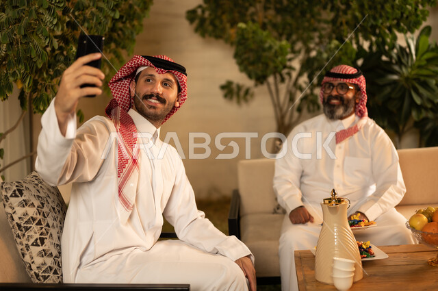 Taking a selfie with your mobile phone, chatting, a happy family gathering, expressions of affection and love, enjoying time with your children, an elderly Saudi Arabian Gulf man wearing a traditional thobe and shemagh sitting on the sofa next to his young son in the living room