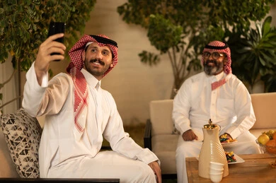 التقاط صورة سيلفي تذكارية عبر الهاتف المحمول، التسامر وتبادل الاحاديث، جلسة عائلية سعيدة، تعابير المودة والمحبة، الاستمتاع بقضاء وقت مع الابناء، رجل كبير بالسن عربي خليجي سعودي يرتدي الثوب التقليدي والشماغ يجلس على الاريكة بجانب ابنه الشاب في غرفة الجلوس