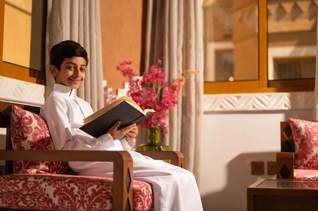 اكتساب مصطلحات وأفكار ثقافية مميزة، القراءة والمطالعة عادة يومية، الشعور بالمتعة والتسلية، طفل عربي خليجي سعودي يرتدي الزي التقليدي ثوب أبيض يجلس في غرفة تراثية ذات تصميم مزخرف وعصري يحمل بيده كتاب وينظر إلى الكاميرا، الاستمتاع في قضاء الأوقات الترفيهية