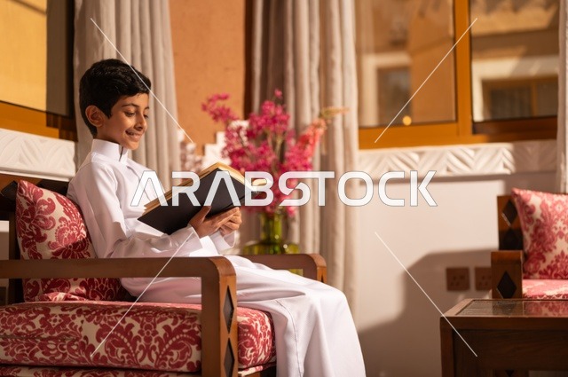 القراءة والمطالعة عادة يومية، اكتساب مصطلحات وأفكار ثقافية مميزة، الشعور بالمتعة والتسلية، طفل عربي خليجي سعودي يرتدي الزي التقليدي ثوب أبيض يجلس في غرفة تراثية ذات تصميم مزخرف وعصري يحمل بيده كتاب، الاستمتاع في قضاء الأوقات الترفيهية