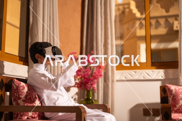 الشعور بالمتعة والتسلية بمحاكاة العالم الخيالي، استخدام نظارات الواقع الافتراضي, تقنيات وأجهزة حديثة،  طفل عربي خليجي سعودي يرتدي الزي التقليدي ثوب أبيض يجلس في غرفة تراثية ذات تصميم مزخرف وعصري، الاستمتاع في قضاء الأوقات الترفيهية
