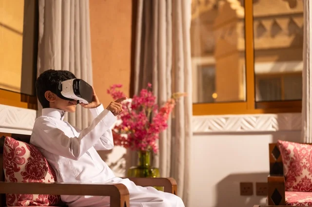 استخدام نظارات الواقع الافتراضي, تقنيات وأجهزة حديثة، الشعور بالمتعة والتسلية بمحاكاة العالم الخيالي، طفل عربي خليجي سعودي يرتدي الزي التقليدي ثوب أبيض يجلس في غرفة تراثية ذات تصميم مزخرف وعصري  ويشير إلى شيء، الاستمتاع في قضاء الأوقات الترفيهية