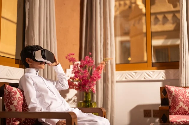 الشعور بالمتعة والتسلية بمحاكاة العالم الخيالي، استخدام نظارات الواقع الافتراضي, تقنيات وأجهزة حديثة،  طفل عربي خليجي سعودي يرتدي الزي التقليدي ثوب أبيض يجلس في غرفة تراثية ذات تصميم مزخرف وعصري، الاستمتاع في قضاء الأوقات الترفيهية