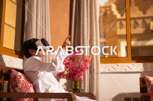 استخدام نظارات الواقع الافتراضي, تقنيات وأجهزة حديثة، الشعور بالمتعة والتسلية بمحاكاة العالم الخيالي، طفل عربي خليجي سعودي يرتدي الزي التقليدي ثوب أبيض يجلس في غرفة تراثية ذات تصميم مزخرف وعصري  ويشير إلى شيء، الاستمتاع في قضاء الأوقات الترفيهية