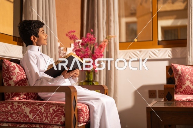 القراءة والمطالعة عادة يومية، اكتساب مصطلحات وأفكار ثقافية مميزة، الشعور بالمتعة والتسلية، طفل عربي خليجي سعودي يرتدي الزي التقليدي ثوب أبيض يجلس في غرفة تراثية ذات تصميم مزخرف وعصري يحمل بيده كتاب، الاستمتاع في قضاء الأوقات الترفيهية