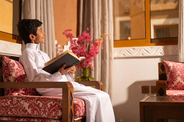 القراءة والمطالعة عادة يومية، اكتساب مصطلحات وأفكار ثقافية مميزة، الشعور بالمتعة والتسلية، طفل عربي خليجي سعودي يرتدي الزي التقليدي ثوب أبيض يجلس في غرفة تراثية ذات تصميم مزخرف وعصري يحمل بيده كتاب، الاستمتاع في قضاء الأوقات الترفيهية