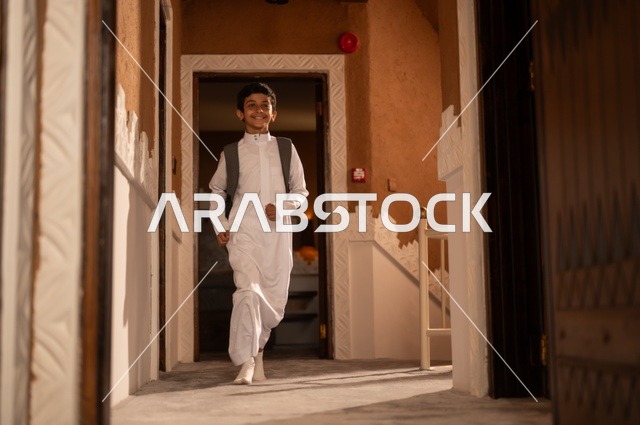 Preparing for the start of a new school day, striving for excellence and success, getting ready and preparing to go to school, an Arab Gulf Saudi child wearing traditional attire, a white thobe, carrying a school bag on his back and looking at something, full body, background of a house door.