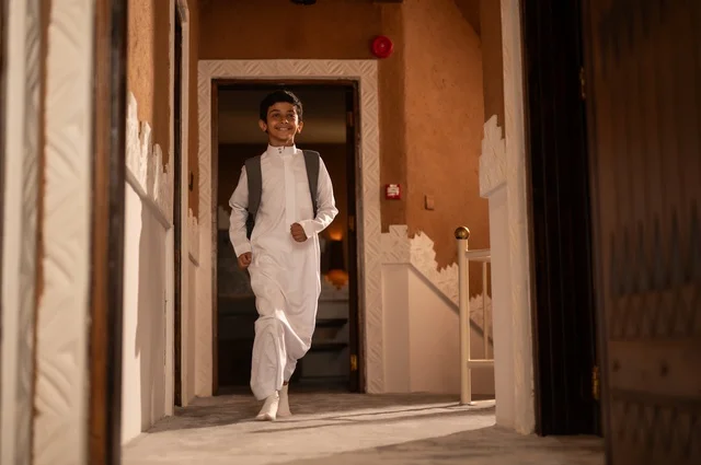 التحضير لبداية يوم دراسي جديد, الحرص على التفوق والنجاح، الاستعداد والتجهز للذهاب الى المدرسة, طفل عربي خليجي سعودي يرتدي الزي التقليدي ثوب أبيض يحمل على ظهره حقيبة مدرسية وتنظر إلى شيء ما, الجسم بالطول الكامل, خلفية باب المنزل