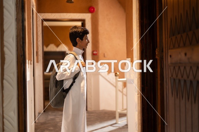 الحرص على التفوق والنجاح، التحضير لبداية يوم دراسي جديد,  الاستعداد والتجهز للذهاب الى المدرسة, طفل عربي خليجي سعودي يرتدي الزي التقليدي ثوب أبيض يحمل على ظهره حقيبة مدرسية وينظر إلى شيء ما,  خلفية باب المنزل