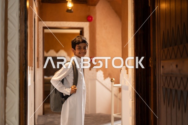 Getting ready and preparing to go to school, looking at the camera with a smile and innocence, striving for excellence and success, preparing for the start of a new school day, an Arab Gulf Saudi child wearing traditional attire, a white thobe, carrying a school bag on his back, with the background of a home door.