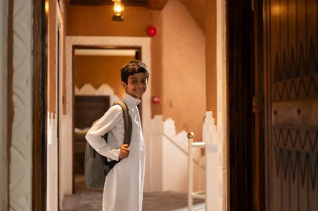 الاستعداد والتجهز للذهاب الى المدرسة, النظر إلى الكاميرا بابتسامة وبراءة، الحرص على التفوق والنجاح، التحضير لبداية يوم دراسي جديد, طفل عربي خليجي سعودي يرتدي الزي التقليدي ثوب أبيض يحمل على ظهره حقيبة مدرسية,  خلفية باب المنزل