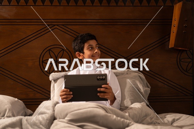 Getting ready for bedtime, using technology in daily life, browsing social media, reviewing lessons and information before sleep, an Arab Gulf Saudi child wearing traditional attire, a white thobe, lying on a clean bed and using a tablet device.
