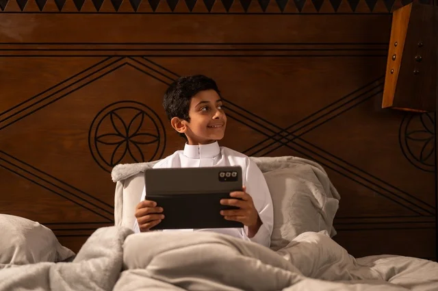 الاستعداد لوقت النوم, استخدام التكنولوجيا في الحياة اليومية، تصفح مواقع التواصل الاجتماعي، مراجعة الدروس والمعلومات قبل النوم, طفل عربي خليجي سعودي يرتدي الزي التقليدي ثوب أبيض يستلقي على سرير نظيف ويستخدم التابلت الجهاز اللوحي