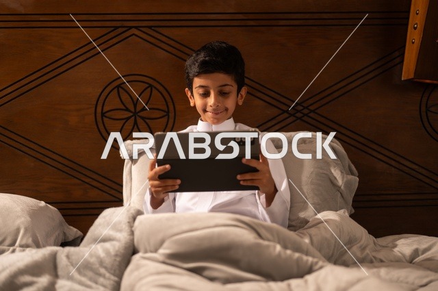 مراجعة الدروس والمعلومات قبل النوم بتقنيات حديثة، استخدام التكنولوجيا في الحياة اليومية، الاستعداد لوقت النوم, تصفح مواقع التواصل الاجتماعي، طفل عربي خليجي سعودي يرتدي الزي التقليدي ثوب أبيض يستلقي على سرير نظيف ويستخدم التابلت الجهاز اللوحي