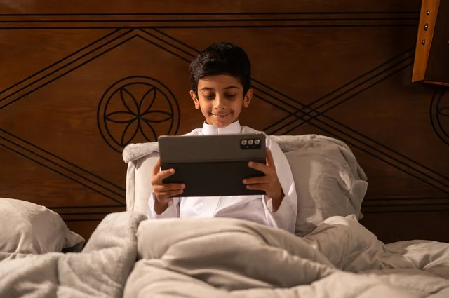 مراجعة الدروس والمعلومات قبل النوم بتقنيات حديثة، استخدام التكنولوجيا في الحياة اليومية، الاستعداد لوقت النوم, تصفح مواقع التواصل الاجتماعي، طفل عربي خليجي سعودي يرتدي الزي التقليدي ثوب أبيض يستلقي على سرير نظيف ويستخدم التابلت الجهاز اللوحي