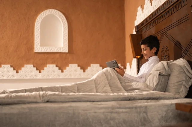 Getting ready for bedtime, using technology in daily life, browsing social media, reviewing lessons and information before sleep, an Arab Gulf Saudi child wearing traditional attire, a white thobe, lying on a clean bed and using a tablet device.
