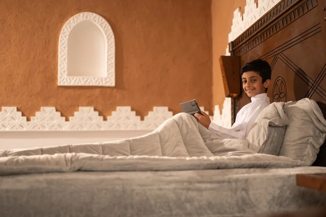 النظر إلى الكاميرا، الاستعداد لوقت النوم, استخدام التكنولوجيا في الحياة اليومية، تصفح مواقع التواصل الاجتماعي، مراجعة الدروس والمعلومات قبل النوم, طفل عربي خليجي سعودي يرتدي الزي التقليدي ثوب أبيض يستلقي على سرير نظيف ويستخدم التابلت الجهاز اللوحي