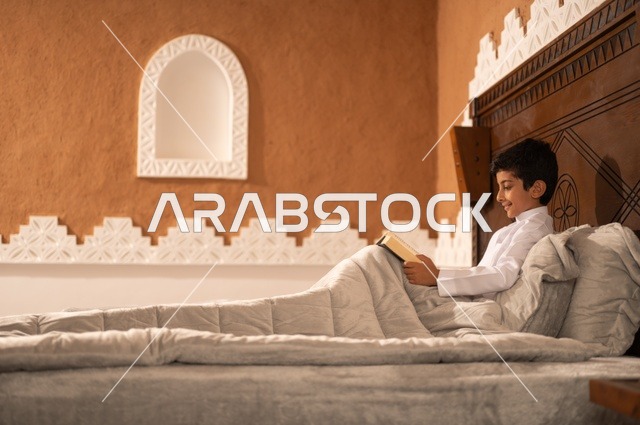 A bedtime story, preparing for sleep and reading evening supplications, the time before sleep, an Arab Gulf Saudi child wearing traditional white attire lying on a bed in a heritage room with a decorative and modern design, holding a book in his hand, reading and acquiring various Arabic terms.