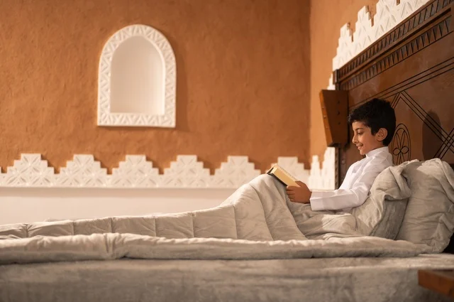 A bedtime story, preparing for sleep and reading evening supplications, the time before sleep, an Arab Gulf Saudi child wearing traditional white attire lying on a bed in a heritage room with a decorative and modern design, holding a book in his hand, reading and acquiring various Arabic terms.