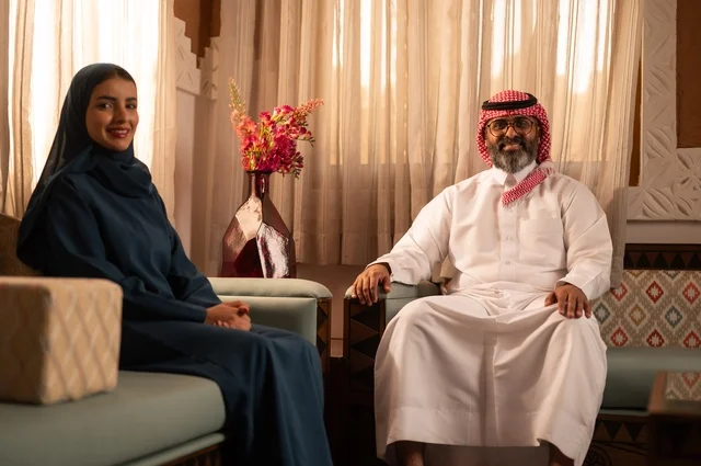 Owning a new house, a Saudi Arabian couple wearing traditional Saudi attire sitting on the sofa and exchanging conversations, the concept of comfort and relaxation, dialogues and discussions between the couple, family stability in the new home, intimacy and love between spouses.