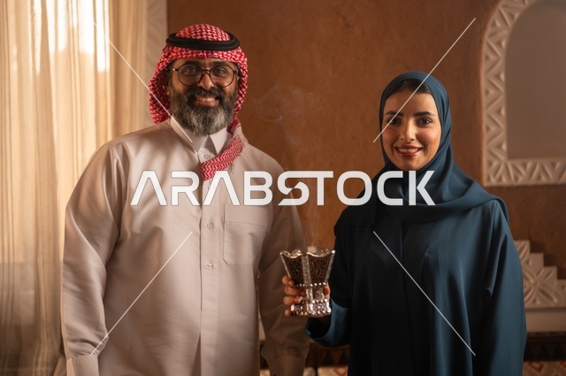 استخدام خشب العود في المناسبات الاجتماعية و الدينية ، التطيب والتعطر  للاستعداد للخروج ، امرأة ترتدي عباءة تحمل حامل البخور( المبخرة )  تقوم بتبخير زوجها، رجل عربي خليجي سعودي يرتدي الزي السعودي يجلس على الاريكة، مفهوم الراحة والاسترخاء، الألفة والمحبة بين الأزواج