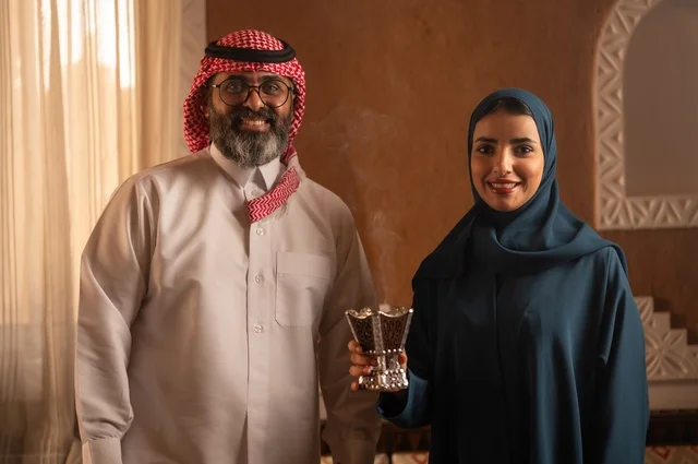 استخدام خشب العود في المناسبات الاجتماعية و الدينية ، التطيب والتعطر  للاستعداد للخروج ، امرأة ترتدي عباءة تحمل حامل البخور( المبخرة )  تقوم بتبخير زوجها، رجل عربي خليجي سعودي يرتدي الزي السعودي يجلس على الاريكة، مفهوم الراحة والاسترخاء، الألفة والمحبة بين الأزواج