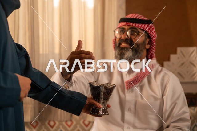 The concept of comfort and relaxation, the use of oud wood on occasions, perfuming and scenting in preparation for going out, a close-up image of a woman wearing an abaya holding a incense holder (mabkhara) as she perfumes her husband, an Arab Gulf Saudi man wearing traditional Saudi attire sitting on the sofa, intimacy and love between spouses.