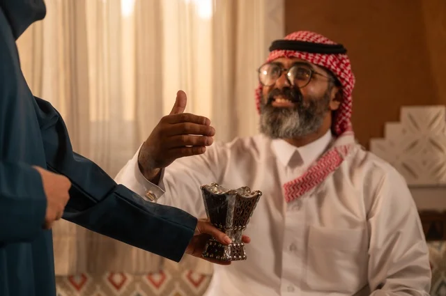 The concept of comfort and relaxation, the use of oud wood on occasions, perfuming and scenting in preparation for going out, a close-up image of a woman wearing an abaya holding a incense holder (mabkhara) as she perfumes her husband, an Arab Gulf Saudi man wearing traditional Saudi attire sitting on the sofa, intimacy and love between spouses.