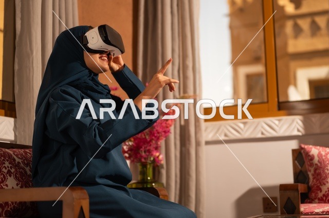 إيماءات التفاعل والاندماج مع الواقع الافتراضي، قضاء أوقات الفراغ بشكل ممتع، امرأة عربية خليجية سعودية ترتدي الحجاب والعباءة تجلس على الاريكة تضع نظارات الفي آر، استخدام الأجهزة التقنية الحديثة والتكنولوجيا في رؤية المشاهد الخيالية
