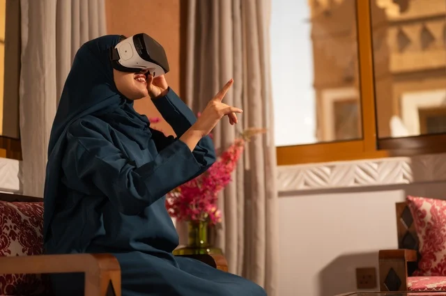 إيماءات التفاعل والاندماج مع الواقع الافتراضي، قضاء أوقات الفراغ بشكل ممتع، امرأة عربية خليجية سعودية ترتدي الحجاب والعباءة تجلس على الاريكة تضع نظارات الفي آر، استخدام الأجهزة التقنية الحديثة والتكنولوجيا في رؤية المشاهد الخيالية